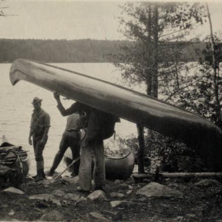 Man holding canoe over his head on the banks of a lake with trees