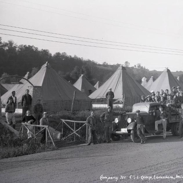 Row of white triangular tents lining a dirt road, with men and trucks lined up in front