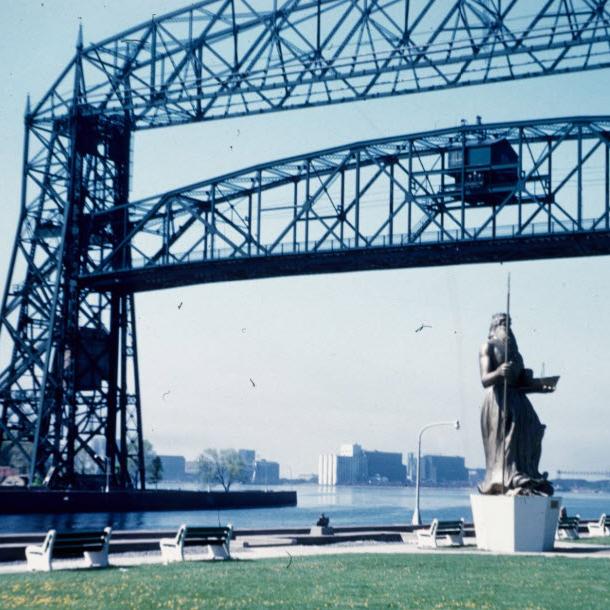 Lift bridge in Duluth silhouetted against a blue sky, with a statue of Neptune holding a trident in the foreground