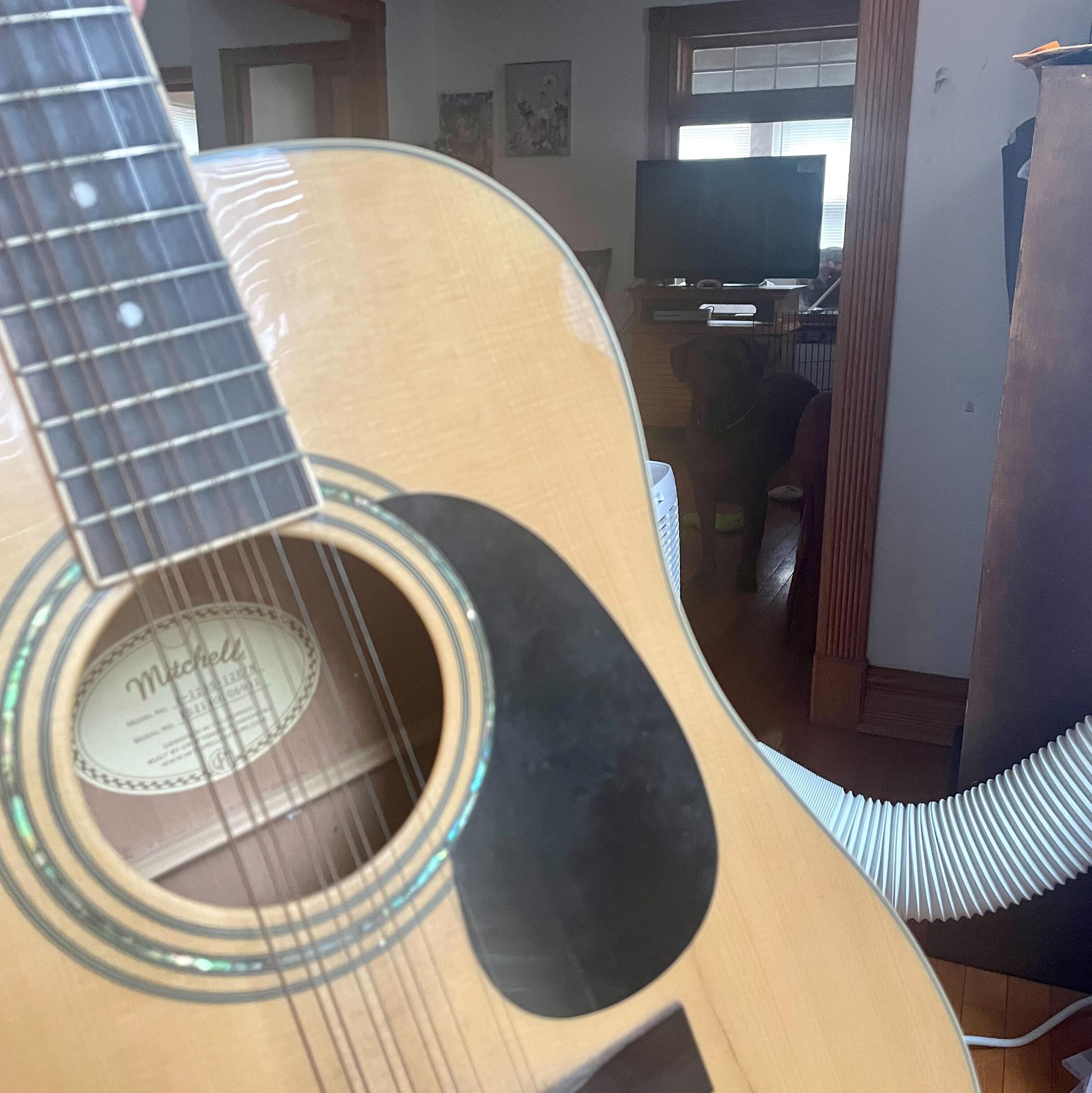 A photo of a wooden 12 string guitar and pick upon a gaming seat. 